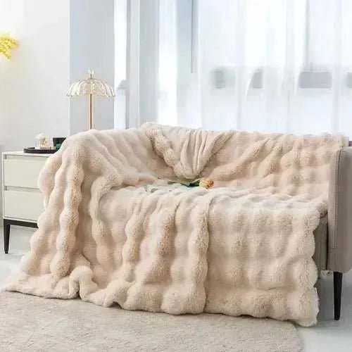 A beige, plush House of Effy TUSCAN THROW is draped over a light-colored sofa next to a white nightstand with a small lamp. The room is well-lit with natural light from a window, evoking winter warmth and cozy comfort.