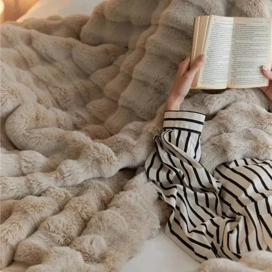 A person in striped pajamas is lying down under the winter warmth of a plush, beige TUSCAN THROW by House of Effy, holding and reading an open book.