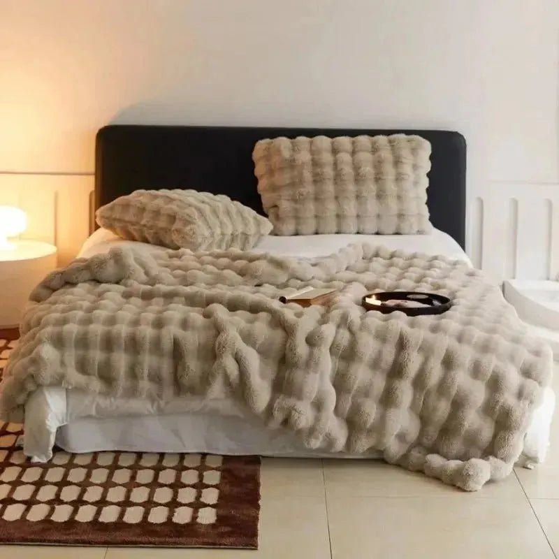 A neatly made bed with a beige, textured bedspread and matching pillows, set against a plain white wall. A lit candle and a book lie on the bed atop a TUSCAN THROW by House of Effy for winter warmth. A lit lamp is seen on the bedside table.