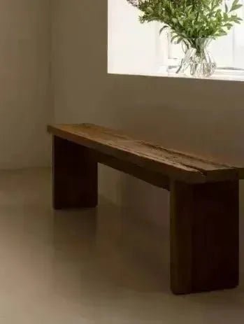 A rustic, minimalist MINIMALIST WOOD BENCH by House of Effy with a natural wood finish sits against a light-colored wall with a small window displaying a glass vase filled with green foliage.