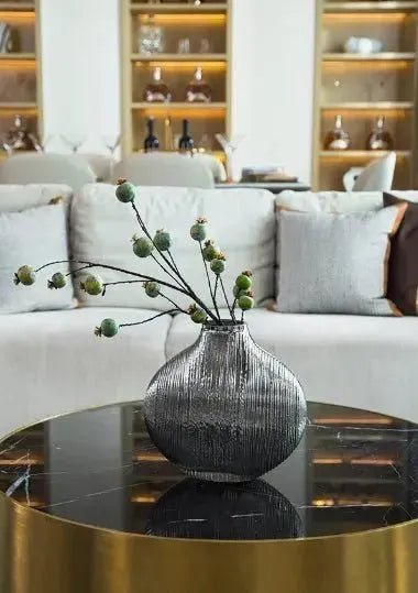 A round House of Effy MERCURY GLASS VASE with dried flowers sits on a black and gold coffee table, reflecting luxury decor in front of a light-colored couch with cushions and a bookshelf in the background.