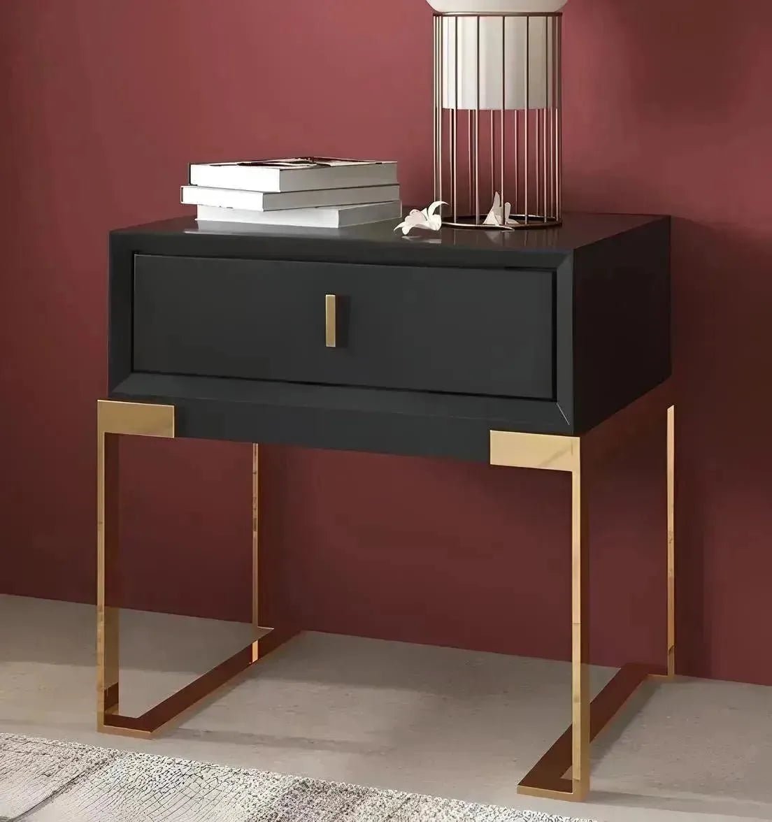 A House of Effy CONTEMPORARY NIGHTSTAND with a drawer, gold metal legs, and a decorative object on top sits against a maroon wall.