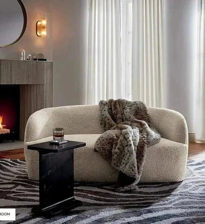 A cozy contemporary living room features a stylish seating area with a House of Effy BOUCLE SOFA, draped with a faux fur blanket, positioned in front of floor-length curtains. A black side table holds a drink, and a lit fireplace is visible in the background.