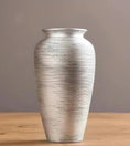 Load image into Gallery viewer, The MODERN CERAMIC VASE from House of Effy, featuring a tall, cylindrical shape with horizontal etched lines, rests on a wooden surface against a plain gray background.
