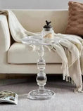 Load image into Gallery viewer, A House of Effy CLEAR ACRYLIC END TABLE showcases modern sophistication beside a beige sofa adorned with a light blanket and textured pillow. On the table rests a decorative jar with a lid, while an open magazine lies invitingly on the floor.
