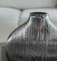 Load image into Gallery viewer, A silver metallic vase with vertical textured lines, reminiscent of a sophisticated MERCURY GLASS VASE by House of Effy, is placed on a reflective surface, with a blurred background of a white sofa.
