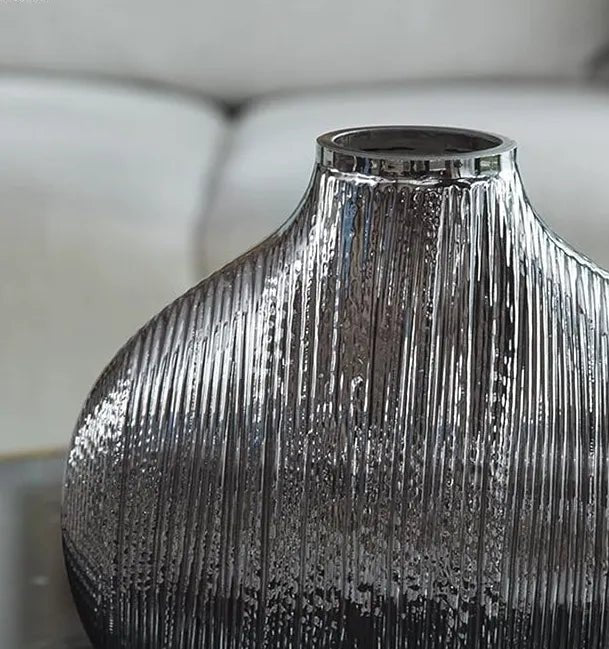 A silver metallic vase with vertical textured lines, reminiscent of a sophisticated MERCURY GLASS VASE by House of Effy, is placed on a reflective surface, with a blurred background of a white sofa.