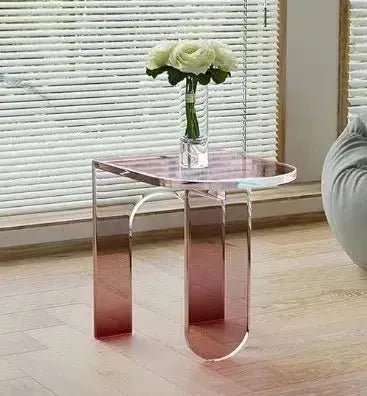 A small, transparent pink acrylic side table with a vase holding three white flowers on top, placed in front of a window with horizontal blinds. This ACYRLIC SIDE TABLE by House of Effy adds a touch of contemporary design to any room.