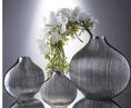 Load image into Gallery viewer, Three ribbed, gray glass vases of varying sizes are displayed on a reflective surface, exuding luxury. One House of Effy MINIMALIST VASE contains white flowers and green foliage.
