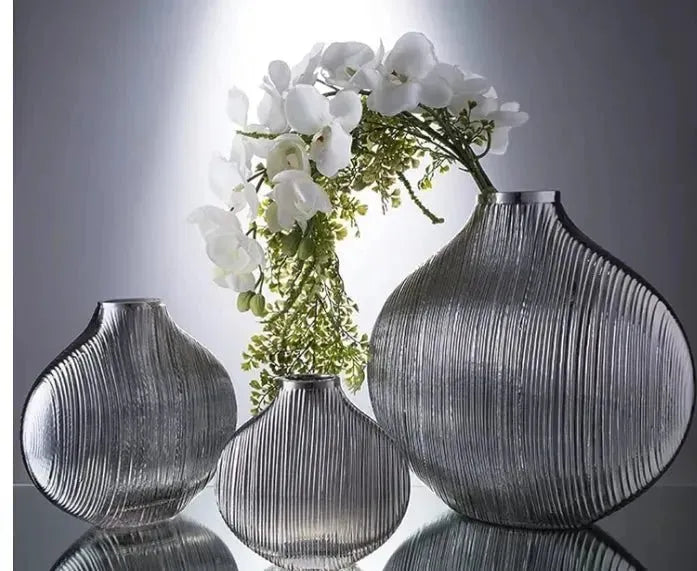 Three ribbed, gray glass vases of varying sizes are displayed on a reflective surface, exuding luxury. One House of Effy MINIMALIST VASE contains white flowers and green foliage.