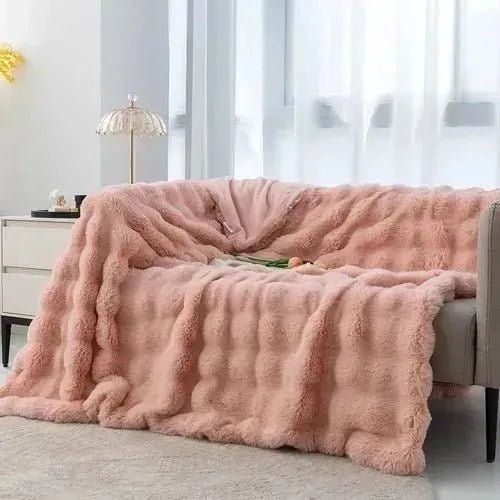 A pink, plush blanket from the House of Effy TUSCAN THROW collection is draped over a gray couch in a bright room with a window, a small side table, and a lamp. Perfect for adding winter warmth to your cozy space.