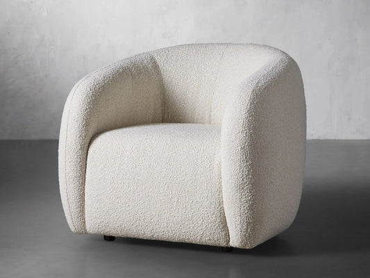 A modern, cream-colored House of Effy BOUCLE SWIVEL CHAIR with a rounded backrest and plush upholstery sits against a gray concrete wall, adding a touch of minimalistic design to the room. Its swivel base enhances both functionality and style.