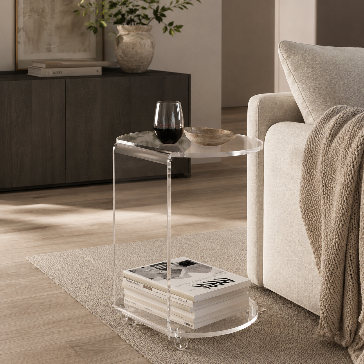 A modern two-tier side table made of transparent acrylic, styled in a living room next to a beige sofa with a knit throw blanket and a glass of red wine on top.