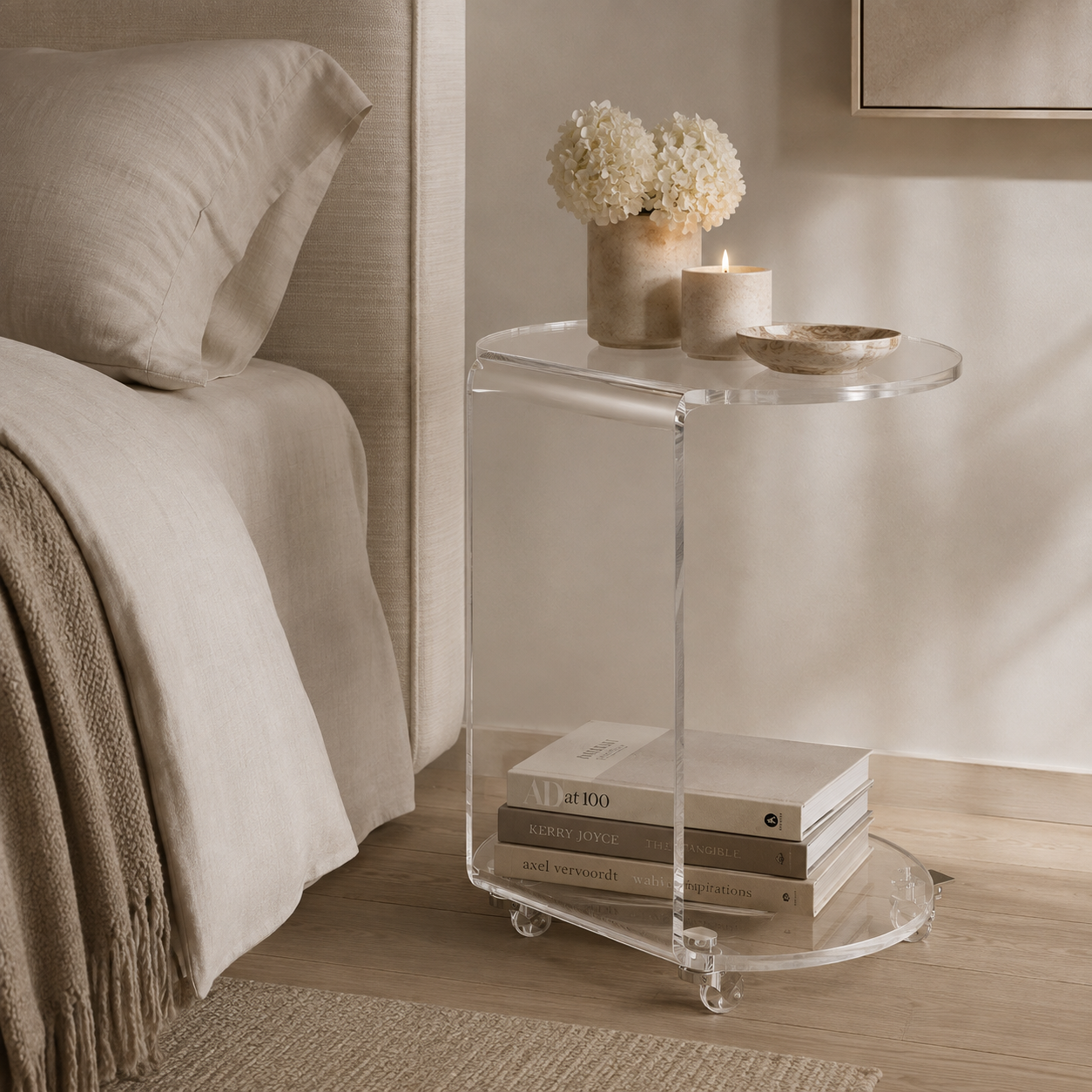 A clear acrylic side table with a rounded rectangular shape, shown in a styled bedroom next to a bed. The table has a top surface and a lower shelf holding books, with decorative items including a vase of white flowers and a candle on top.