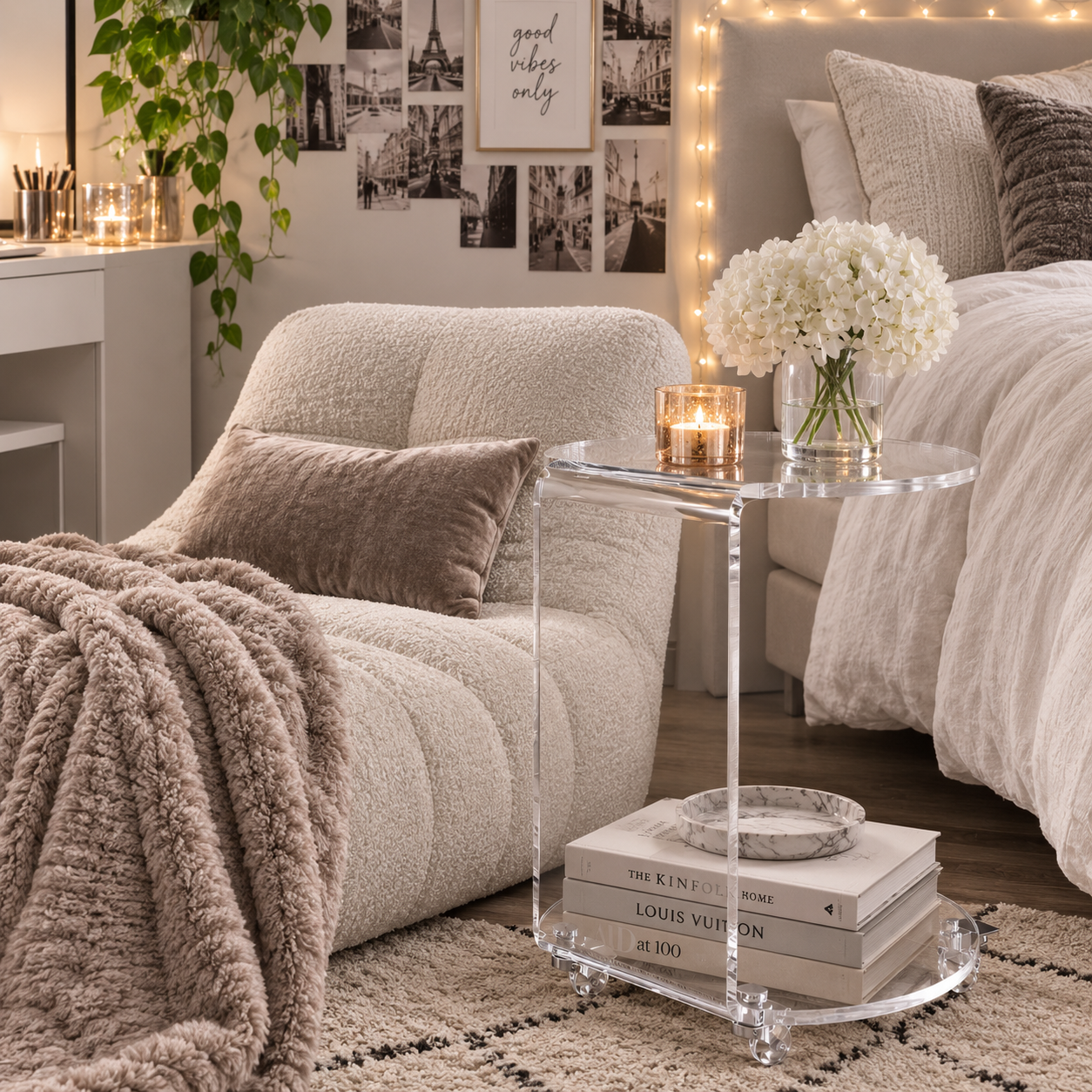 A clear acrylic round side table with a glass top and lower shelf, styled in a cozy bedroom next to a cream bouclé chair and bed. The table holds a lit candle and a stack of books.