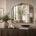 Load image into Gallery viewer, An arched mirror with an ornate crest at the top, mounted on a beige wall above a dark wood console table. The mirror reflects a softly lit living room with a window, curtains, and a fireplace.
