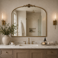 Load image into Gallery viewer, An ornate arched wall mirror with a gold frame hangs above a marble double vanity with a gold faucet in a warm, neutral-toned bathroom.
