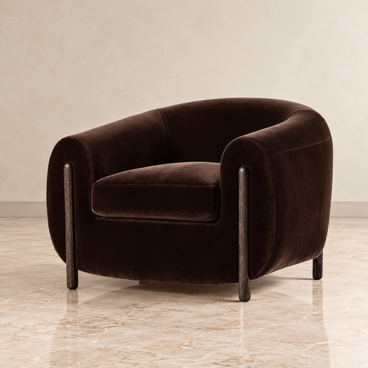 A dark brown velvet barrel chair with a curved backrest and slim dark metal legs, set against a neutral beige wall and light marble floor.