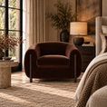 Load image into Gallery viewer, A dark brown velvet barrel chair with dark metal legs, placed in a beautifully styled bedroom next to a bed with a textured beige blanket, a stone side table, and a lit table lamp.
