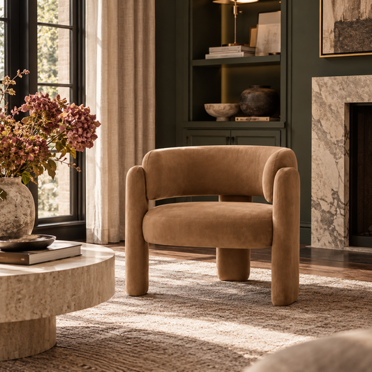 A modern tan velvet armchair with chunky cylindrical legs and curved arms, styled in a luxurious living room next to a stone coffee table and a dark green built-in cabinet.