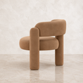 Load image into Gallery viewer, A modern upholstered accent chair in a warm camel brown suede-like fabric, featuring a rounded backrest, thick cylindrical legs, and integrated armrests, set against a neutral textured beige wall and floor.
