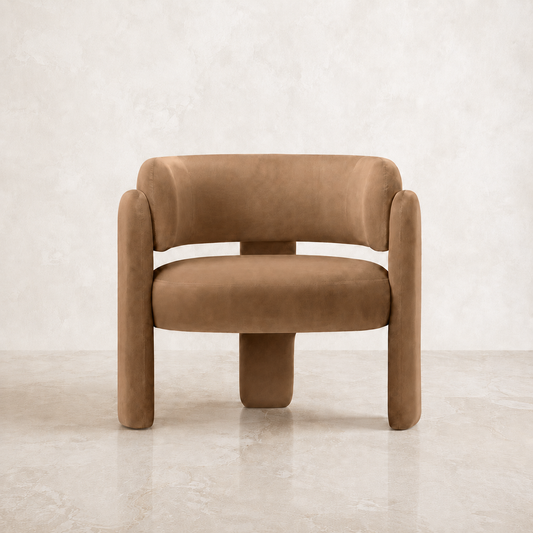 A modern low-profile barrel chair upholstered in a warm camel brown velvet fabric, set against a neutral textured beige wall and floor.