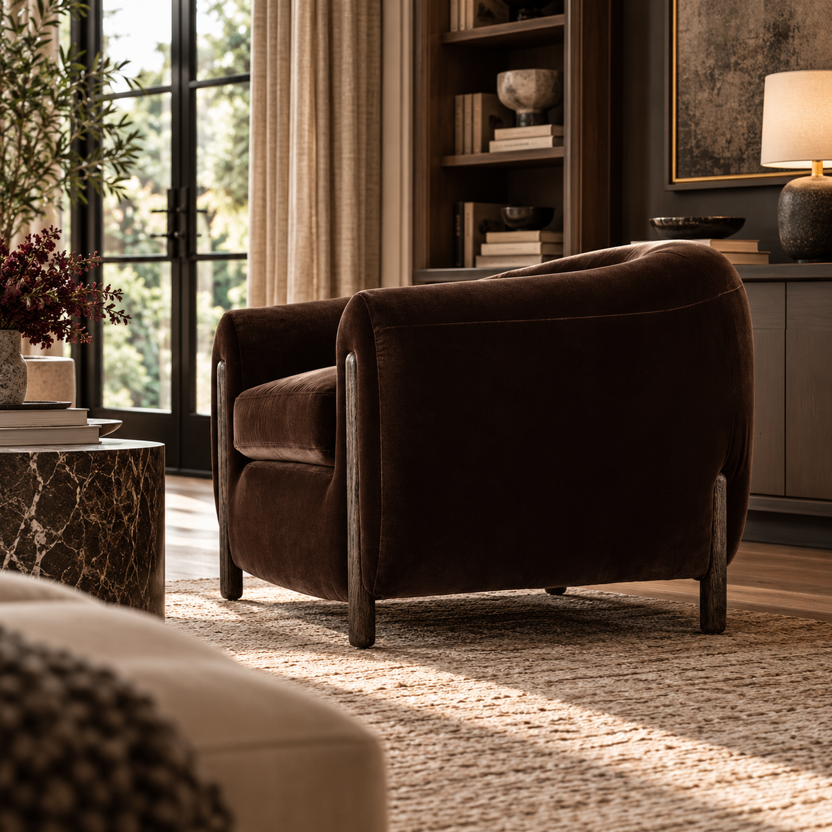 A dark brown velvet barrel chair with dark wood legs, styled in a warm, sunlit living room next to a marble side table and a bookshelf.
