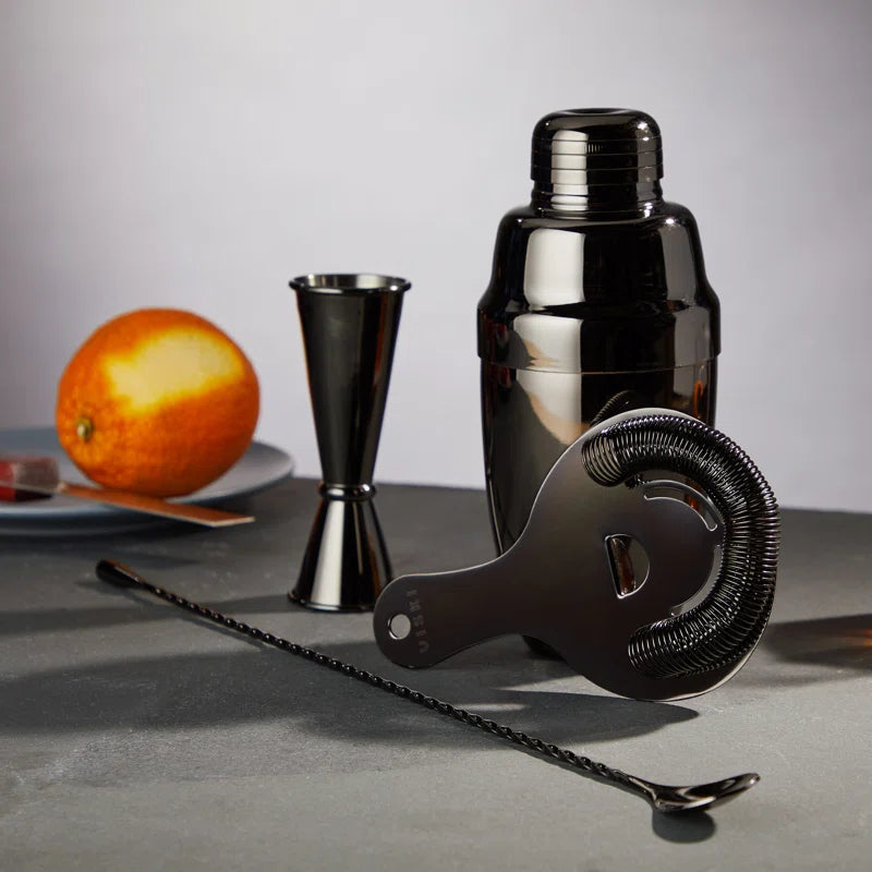 barware set, including a cocktail shaker, jigger, and strainer, on a gray surface with an orange in the background.