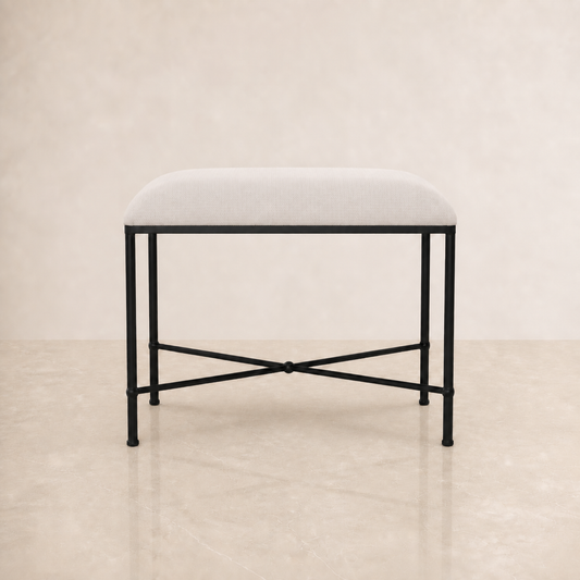 A modern bedroom bench with a light beige upholstered cushion and a black metal frame with an X-shaped base, set against a neutral beige background.