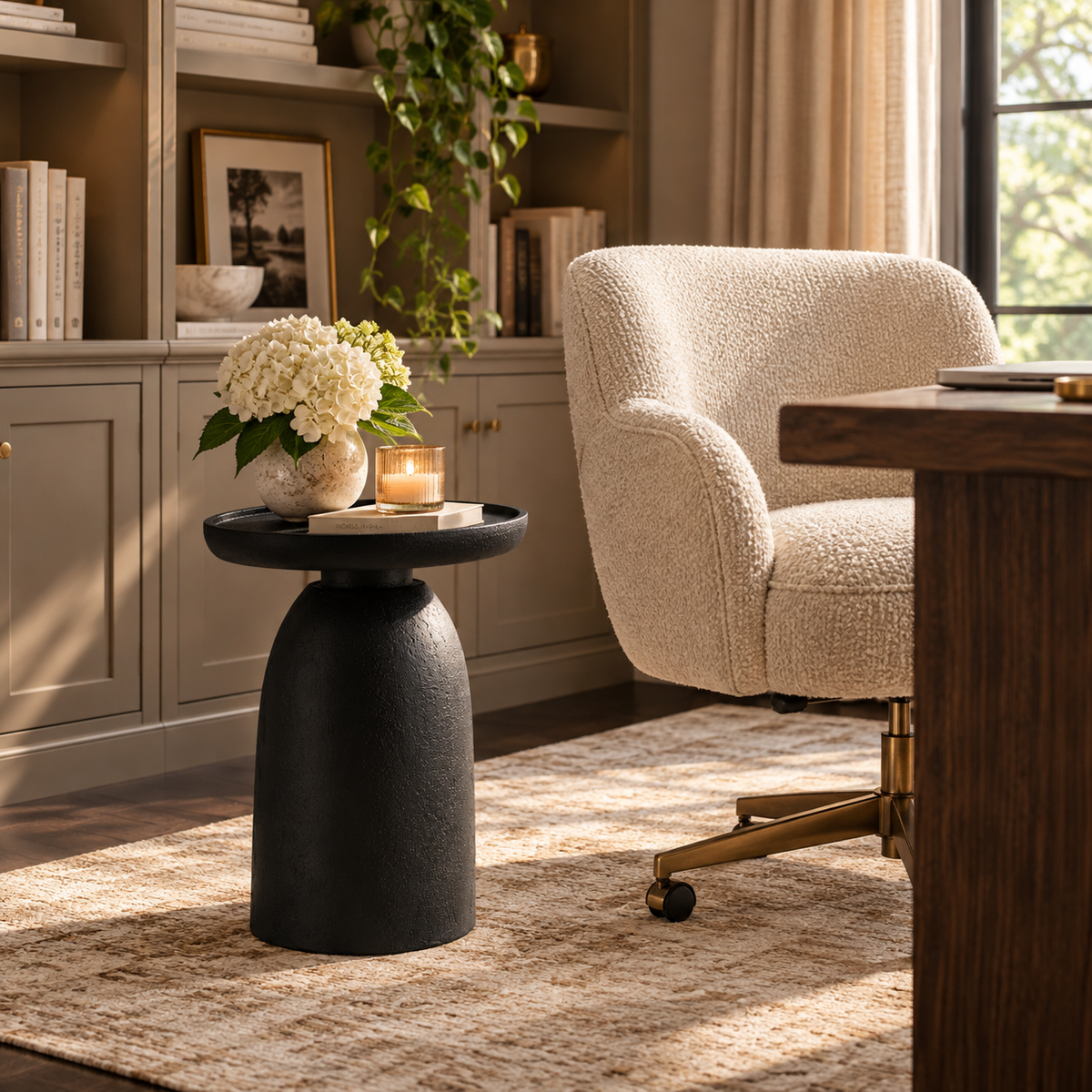 A modern black round side table with a textured, tapered pedestal base stands in a styled home office or living room setting next to a cream bouclé armchair and a wooden desk.