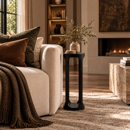 A modern black metal side table with a sculptural curved base stands beside a cream upholstered sofa in a warm, sunlit living room. The table holds a small beige vase with dried branches and a stack of books.