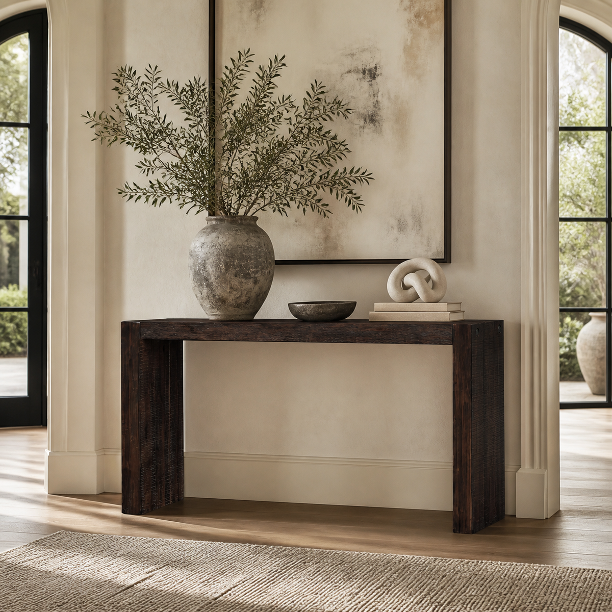 A dark brown console table styled with a large textured gray vase containing green branches, a small dark bowl, and a white sculptural object on books, set against a warm beige wall with a framed abstract print.