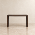 Load image into Gallery viewer, A dark brown wooden console table with a minimalist rectangular design, set against a neutral beige textured wall and marble floor.
