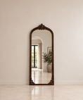 Load image into Gallery viewer, An ornate full-length floor mirror with a dark carved wood frame, leaning against a neutral beige wall in a bright, modern interior.

