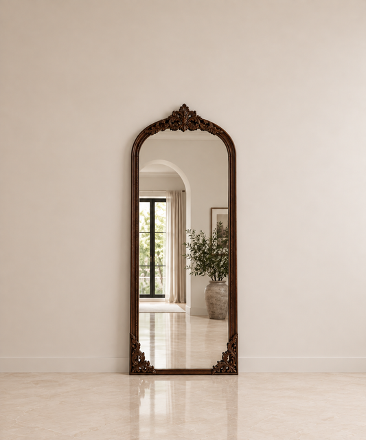 An ornate full-length floor mirror with a dark carved wood frame, leaning against a neutral beige wall in a bright, modern interior.