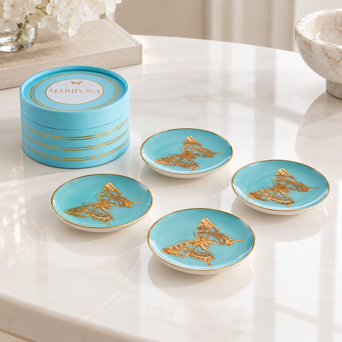 A set of four turquoise blue Mariposa butterfly coasters with gold trim, displayed on a white marble table next to their round turquoise and gold branded storage box.