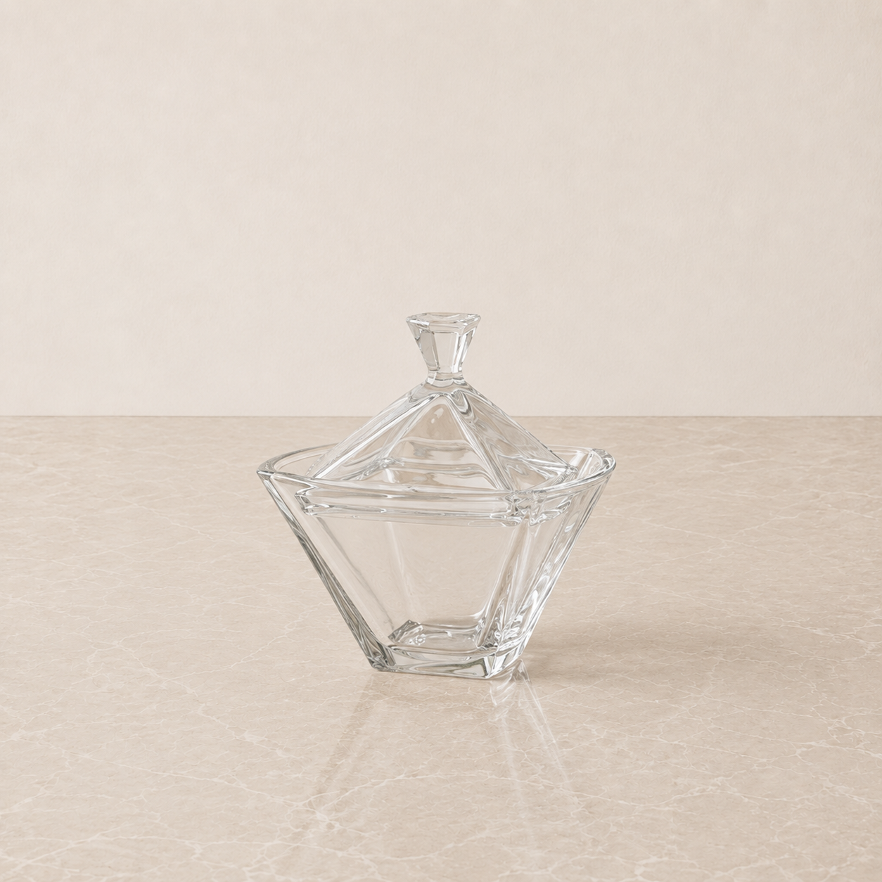 A clear glass lidded candy dish with a faceted geometric design, set on a light beige marble surface against a matching beige background.