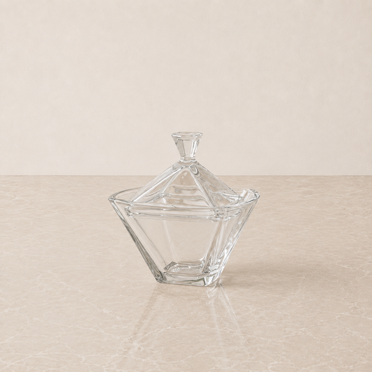 A clear glass lidded candy dish with a faceted geometric design, set on a light beige marble surface against a matching beige background.