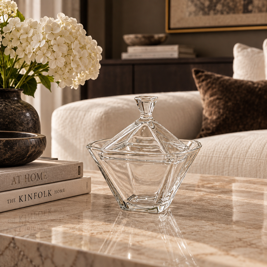 A clear glass lidded bowl with a geometric faceted design sits on a polished marble coffee table in a styled living room setting.