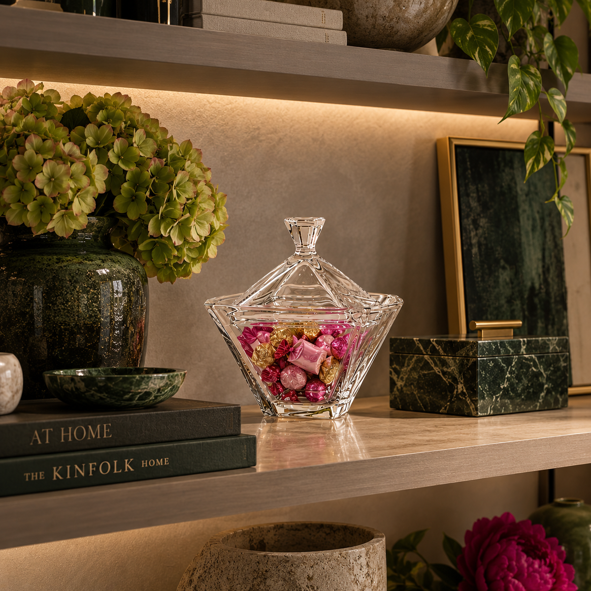 A clear glass lidded candy dish filled with pink dried flowers sits on a wooden shelf beside a green ceramic vase with hydrangeas, books, and framed decor in a styled interior setting.