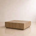 Load image into Gallery viewer, A square wooden coffee table with a light oak finish and visible wood grain, set against a minimalist beige wall and floor.
