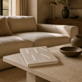 Load image into Gallery viewer, A beige upholstered sofa in a warm, modern living room setting, with a stone coffee table in the foreground holding a book and decorative bowls.

