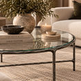Load image into Gallery viewer, A round glass-top coffee table with a distressed dark metal frame, styled in a cozy living room setting with books, a ceramic vase of greenery, a bowl, and candles.
