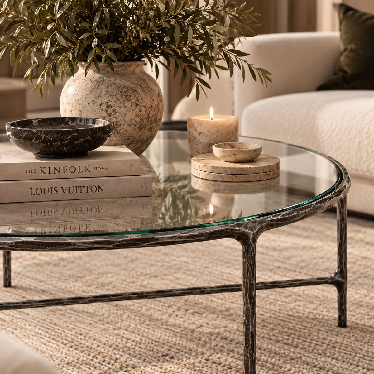 A round glass-top coffee table with a distressed dark metal frame, styled in a cozy living room setting with books, a ceramic vase of greenery, a bowl, and candles.