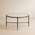 Load image into Gallery viewer, A round coffee table with a clear glass top and a textured dark gray metal frame, set against a neutral beige background.

