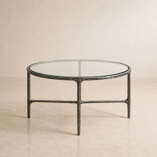 A round coffee table with a clear glass top and a textured dark gray metal frame, set against a neutral beige background.