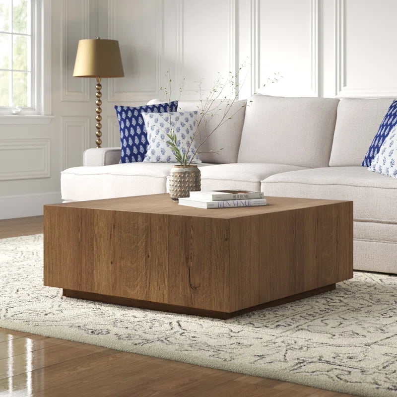 Square wooden coffee table with rustic finish on patterned rug in bright living room with beige sofa and blue pillows