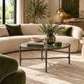 Load image into Gallery viewer, A round glass-top coffee table with a dark metal frame sits in front of a cream boucle sofa in a styled living room. The table is styled with books, a bowl, and a green glass vase with foliage.
