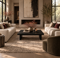 Load image into Gallery viewer, A styled living room interior with two cream sofas, a dark rectangular coffee table, a large textured beige area rug, a black fireplace, and warm natural light streaming through tall windows.

