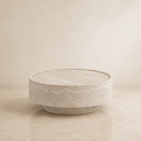 A round, low-profile coffee table with a textured stone or concrete finish in a warm beige-gray color, set against a matching beige background.